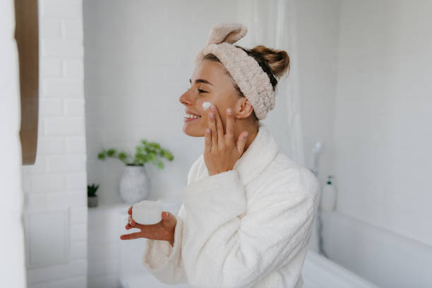 Happy woman in bathrobe applying facial cream in bathroom