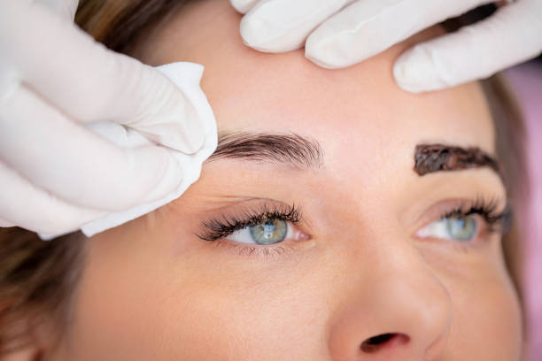 Close-up view of a woman undergoing eyebrow shaping procedure. Two professionals in gloves are touching and examining her eyebrows, which are styled and filled in.