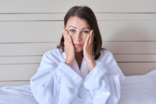 Woman in white robe holding face in hands, looking tired or stressed